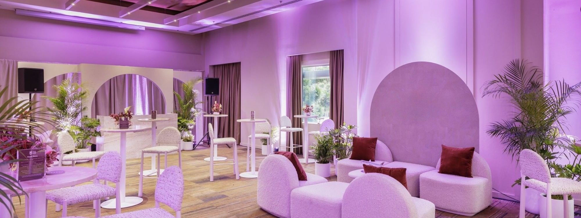Modern event space with purple ambient lighting, white curved seating, potted palms, and hardwood floors