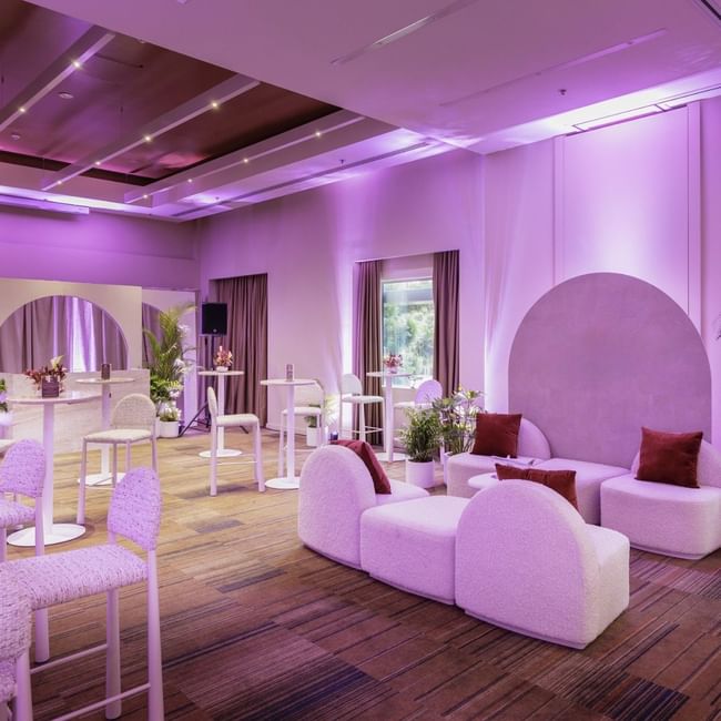 Modern event space with purple ambient lighting, white curved seating, potted palms, and hardwood floors