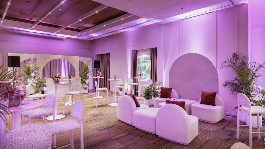 Modern event space with purple ambient lighting, white curved seating, potted palms, and hardwood floors