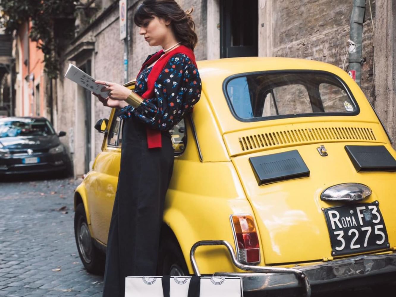 A woman reading a book by a yellow car on a road near Margutta 19