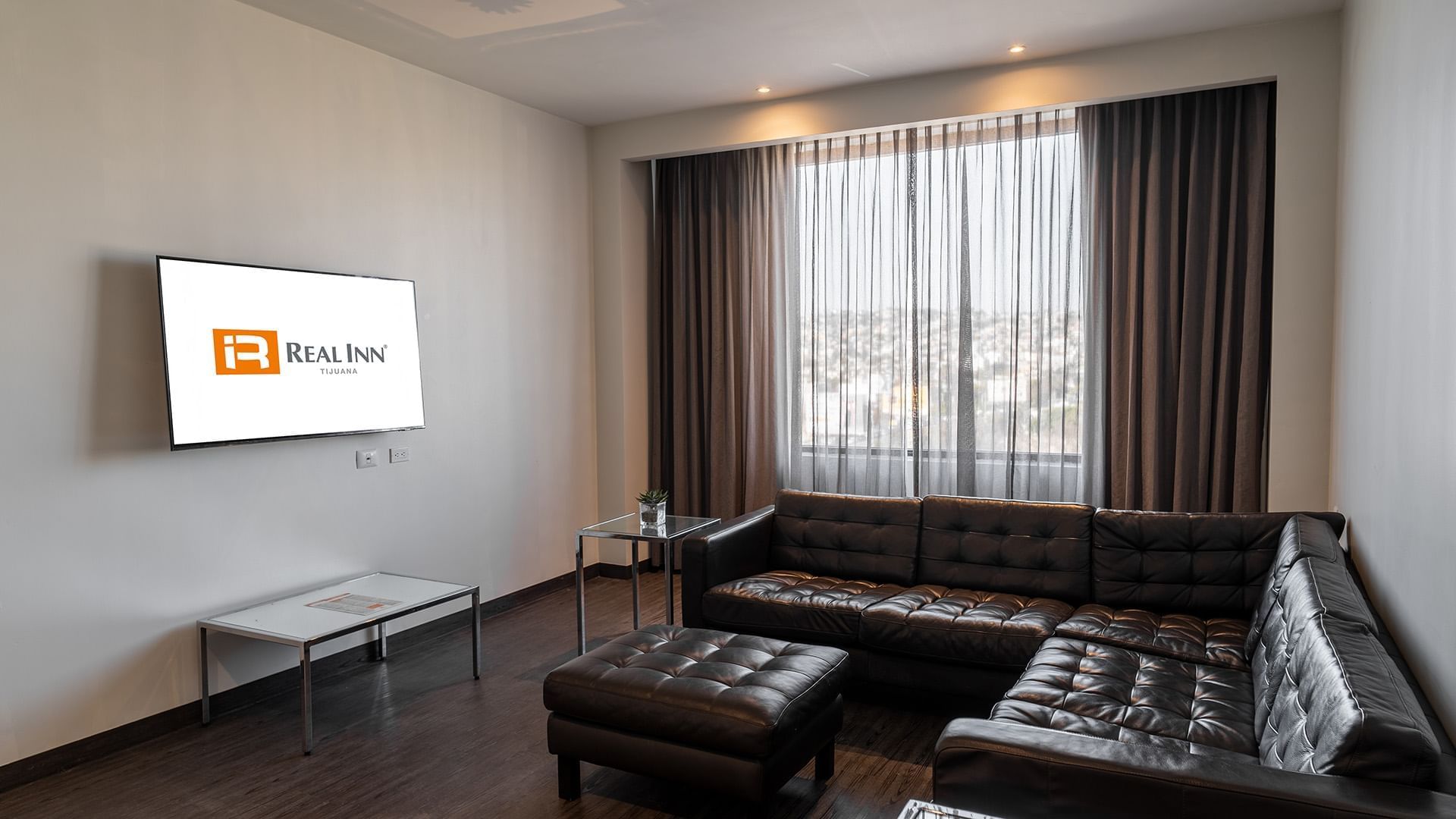 Two Double Master Suite living area with a black tufted leather sofa and city views through curtains at Real Inn Tijuana