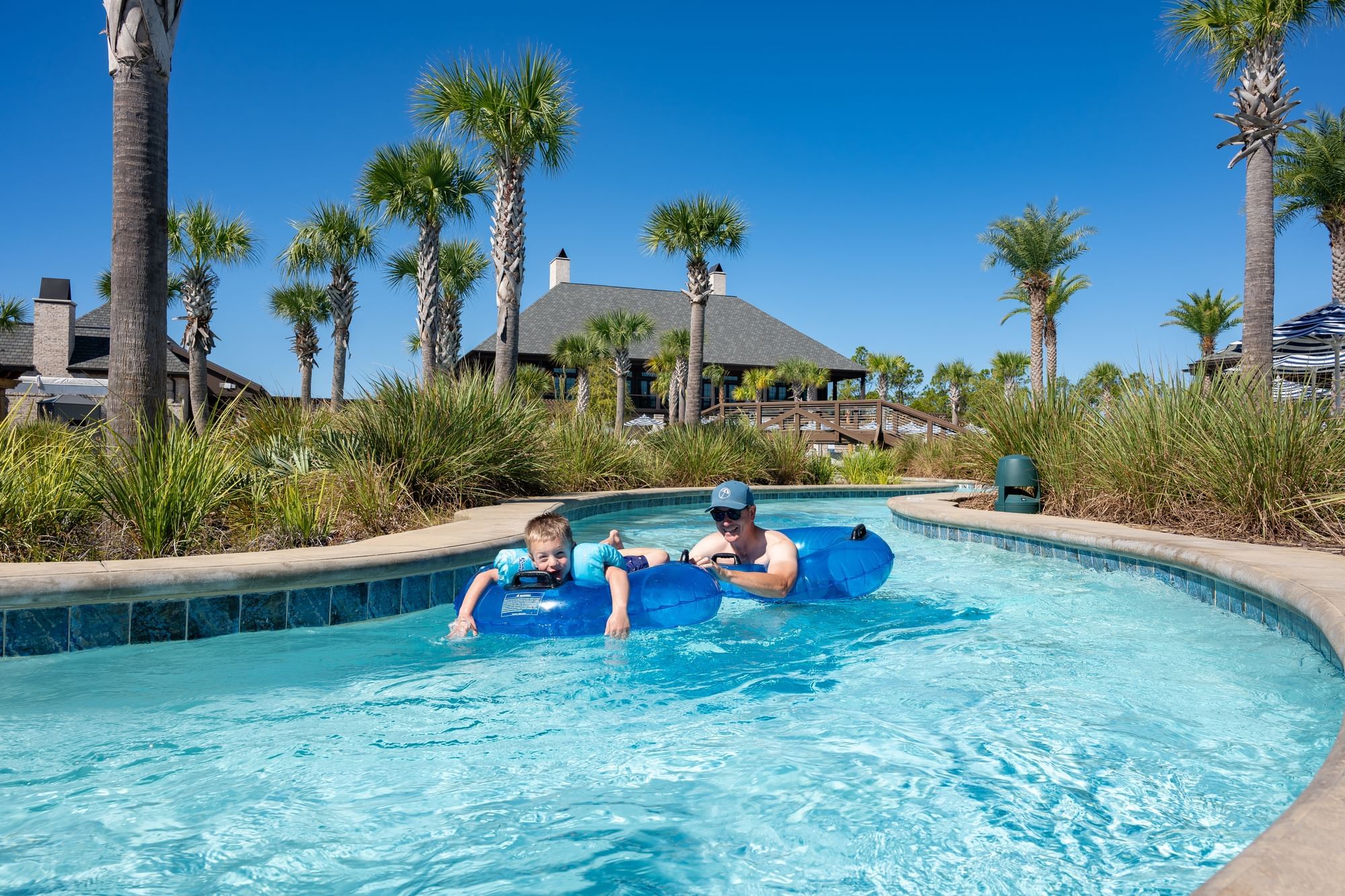 Father and son on tubes in lazy river with a resort and palm trees in background.