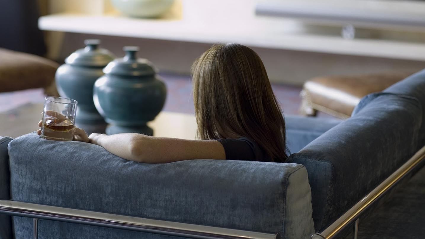 A person with long hair sits on a blue velvet couch holding a drink in The Azure Suite at The May Fair Hotel, London