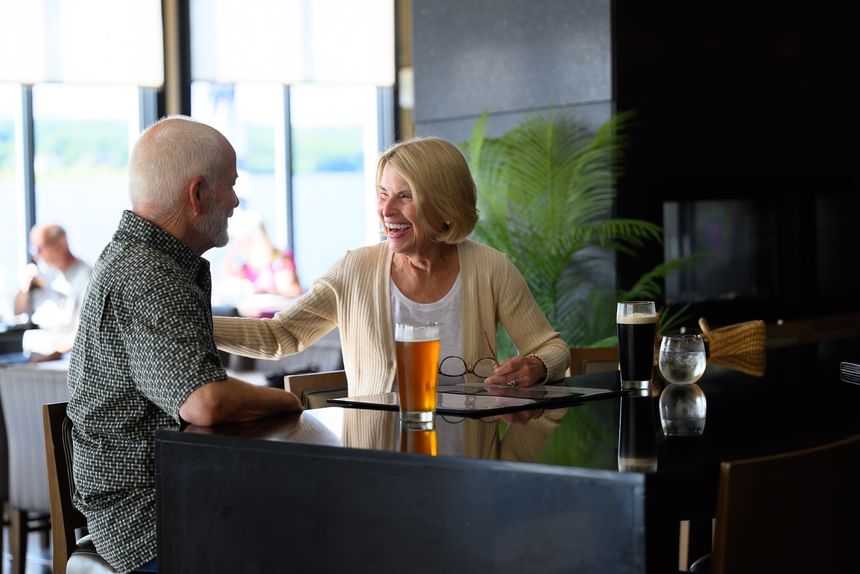 Couple smiling by a glossy black bar under soft lights with drinks in glasses at Rodd Miramichi River