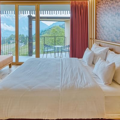 Camera d'albergo con letto matrimoniale, divano letto, tavolo da gioco e balcone con vista sulle montagne.