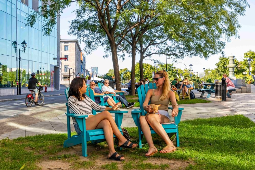 Two women relaxing in turquoise Adirondack chairs in a sunny urban park at Warwick Le Crystal Montreal