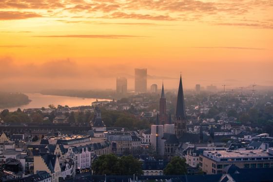 Sonnenuntergang über einer Stadt mit vielen Gebäuden und Türmen, umgeben von Bäumen und einem Gewässer.