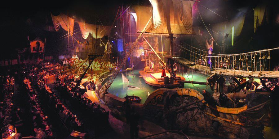 A large indoor pirate-themed dining area with a stage, audience seating, and a large pirate ship in the center of a water feature.