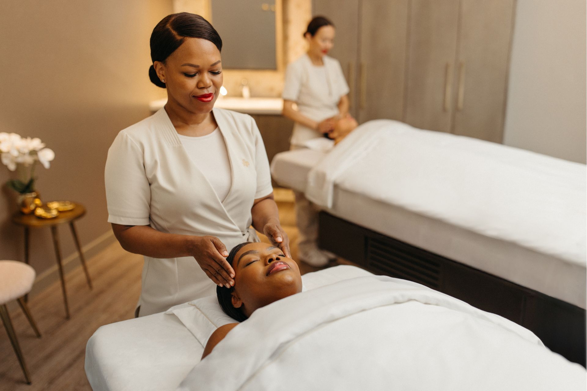 Therapist performing a facial treatment on a client in a spa setting with another client in background.