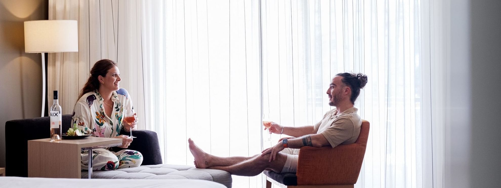 Couple relaxing in hotel room with white curtains, each holding a wine glass, near a bed and a lamp.