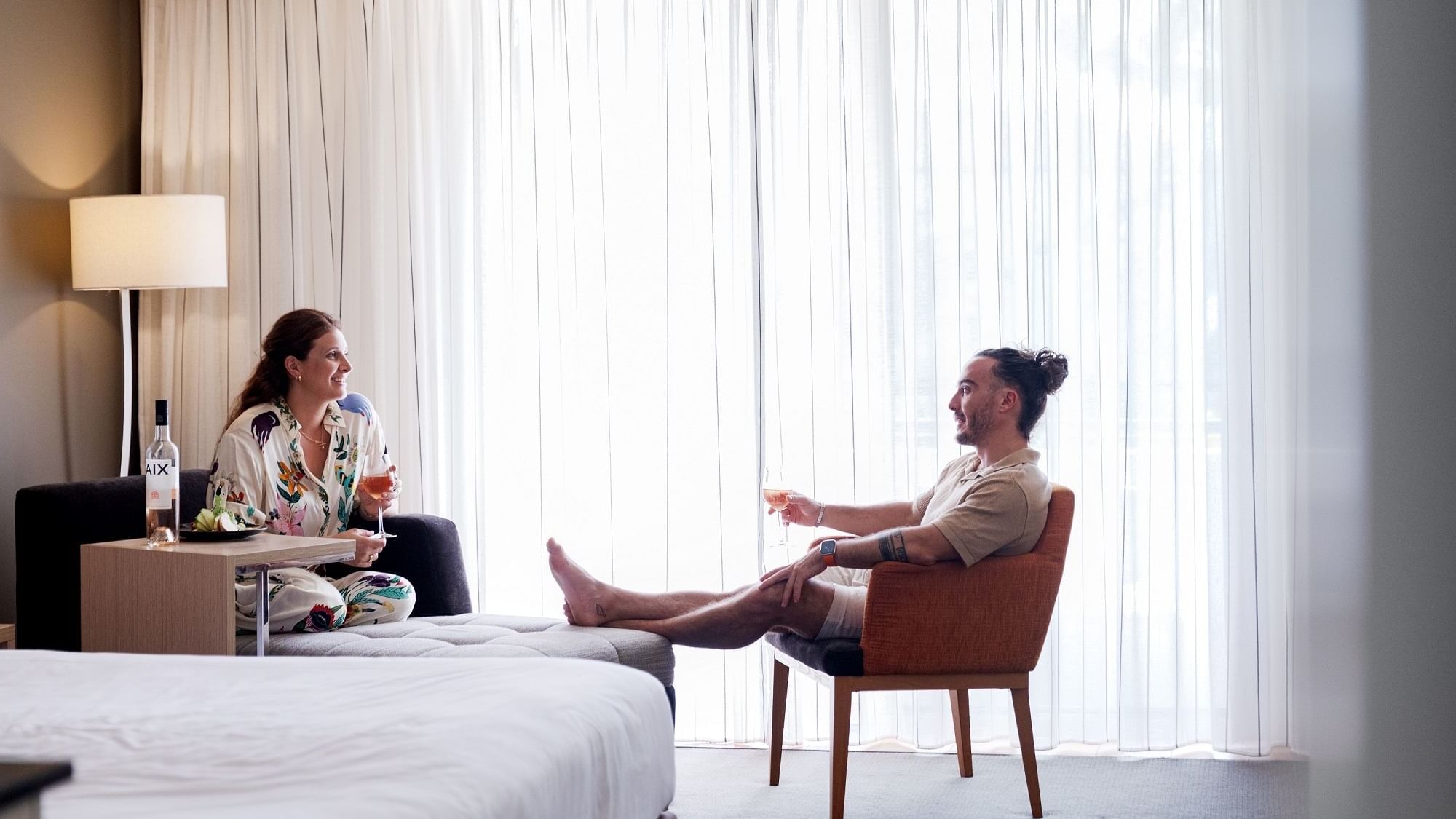 Couple relaxing in hotel room with white curtains, each holding a wine glass, near a bed and a lamp.