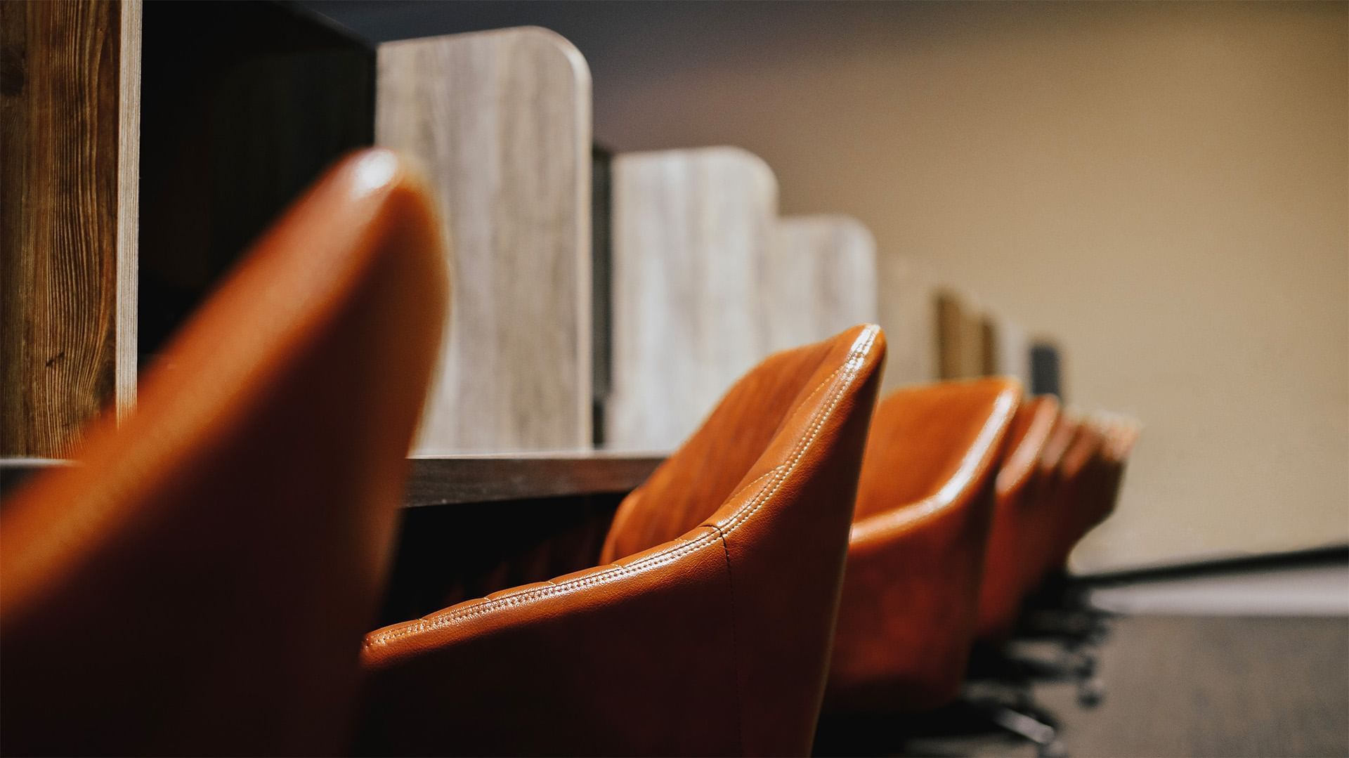 Close-up of tan leather chair backs by wooden privacy dividers along a long counter under soft light at The Brook