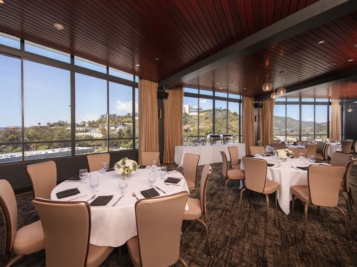 A dining room with round tables set for a meal, large windows, and a wood-paneled ceiling.