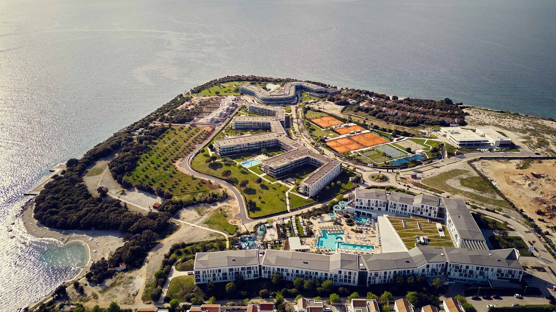 Luftaufnahme eines Küstenresorts mit mehreren Gebäuden, Swimmingpools und Tennisplätzen.