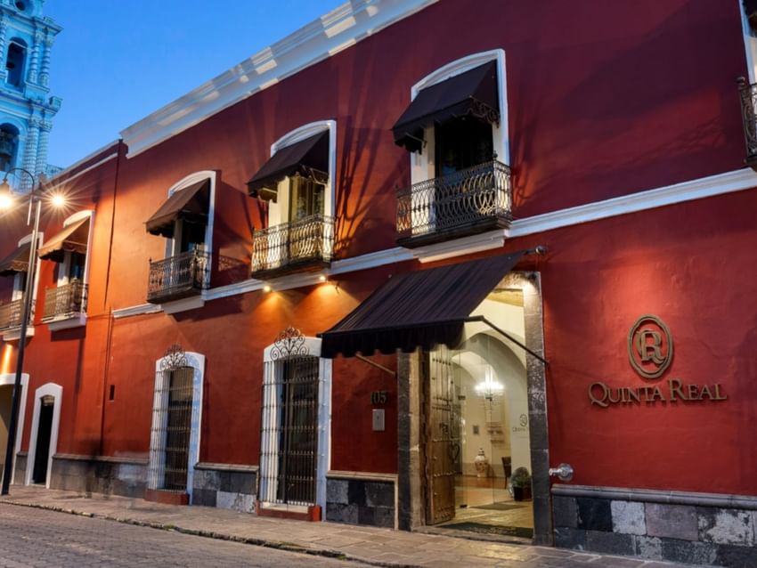 Red colonial facade of Quinta Real Puebla hotel with black awnings and wrought iron balconies at night