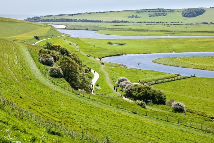 Aerial view of a winding path by a river in lush green countryside with people walking.