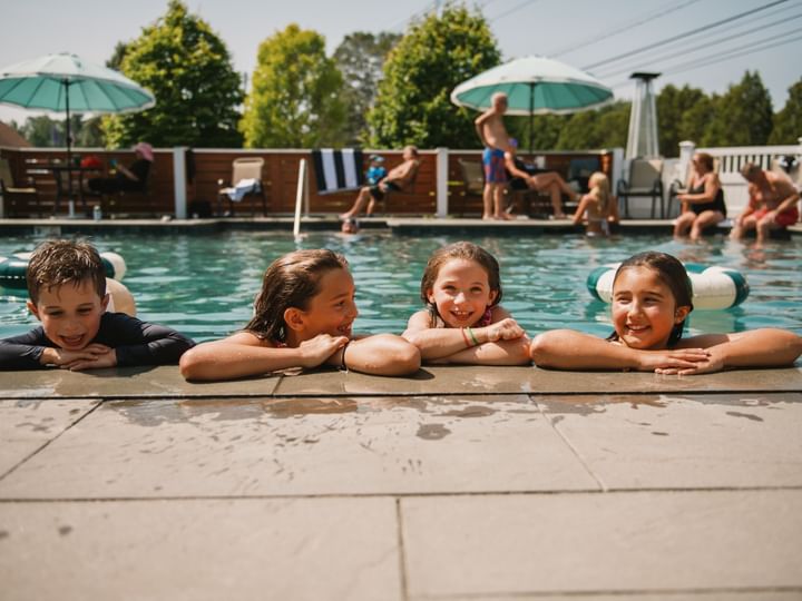Four children leaning on the edge of a swimming pool, smiling at each other.