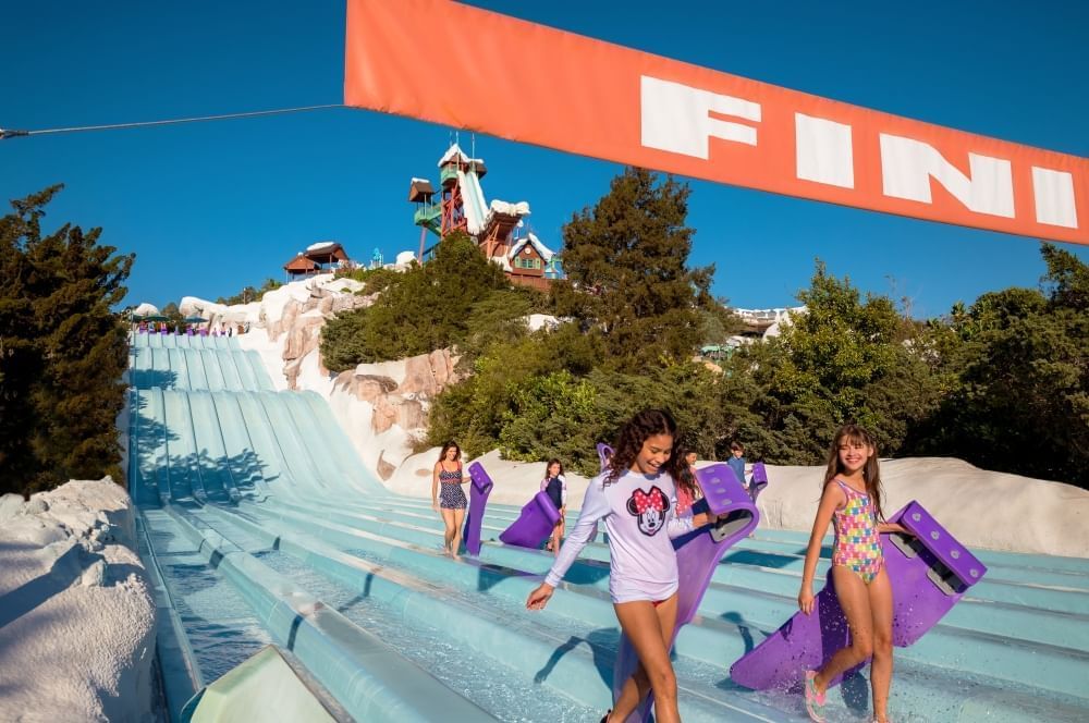 Girls in bathing suits carrying purple mats walk at the base of a multi-lane water slide beneath an orange FINISH sign