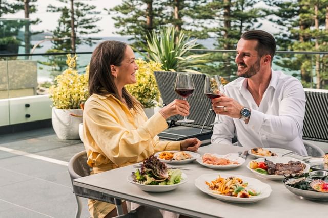 Happy couple toast with red wine glasses by a dining table under tall pine trees surrounding the Elysium Manly scenic coast