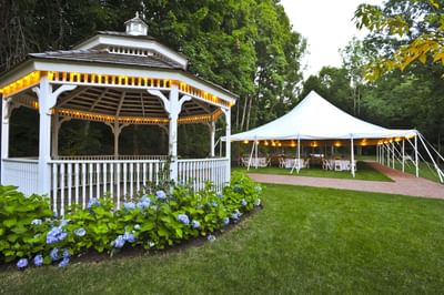 White gazebo and flowers beside a path and a lit white event tent against trees at Avon Old Farms Hotel