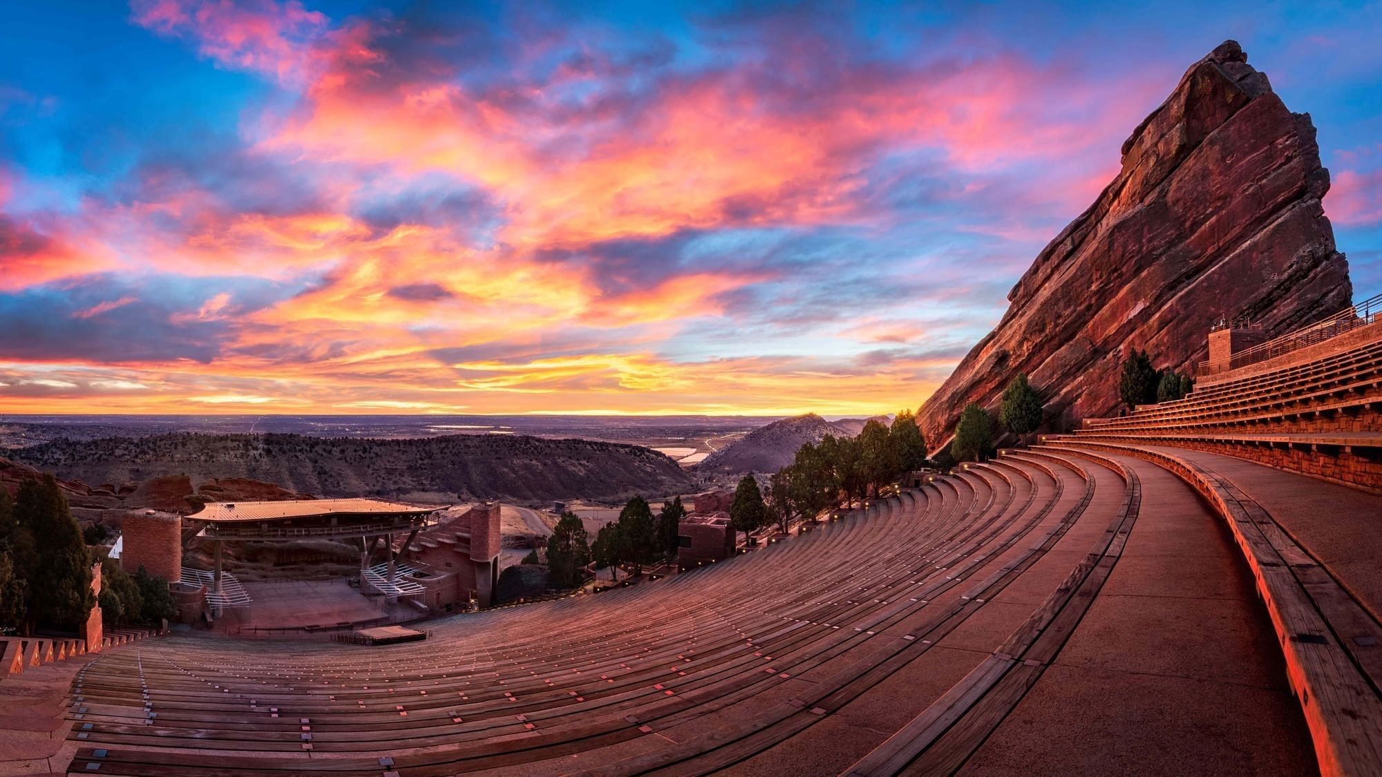 A large outdoor amphitheater with tiered seating, situated on a hillside, under a vibrant sunset sky.
