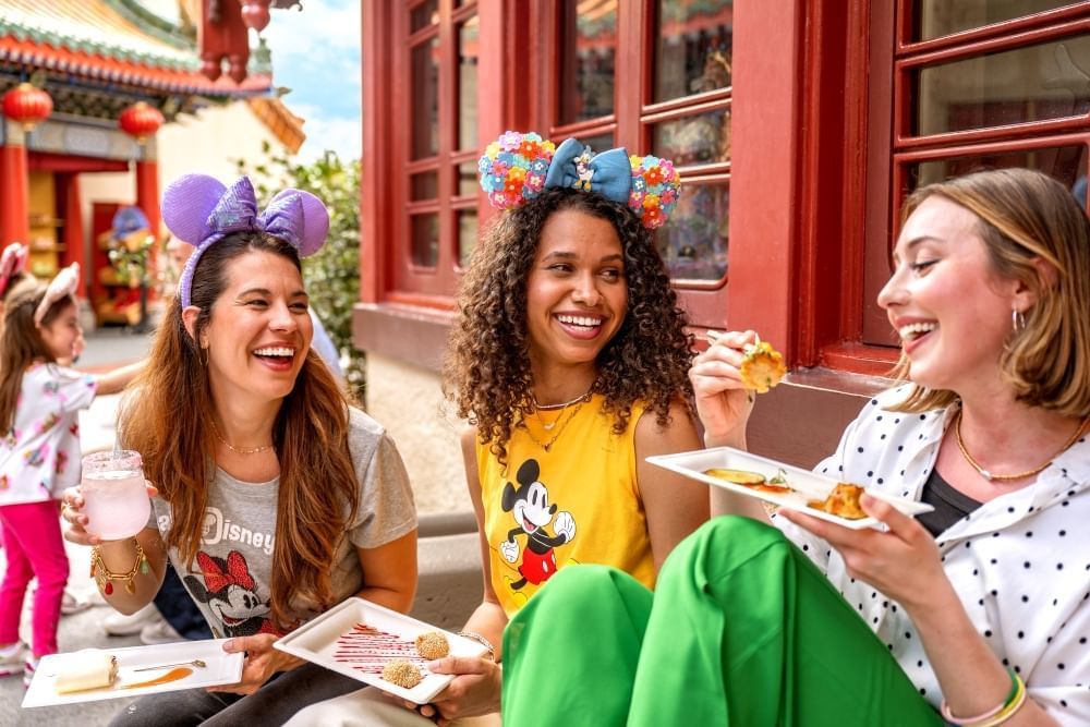 Three women wearing Minnie Mouse ears enjoy plates of food in front of a rust-colored building.