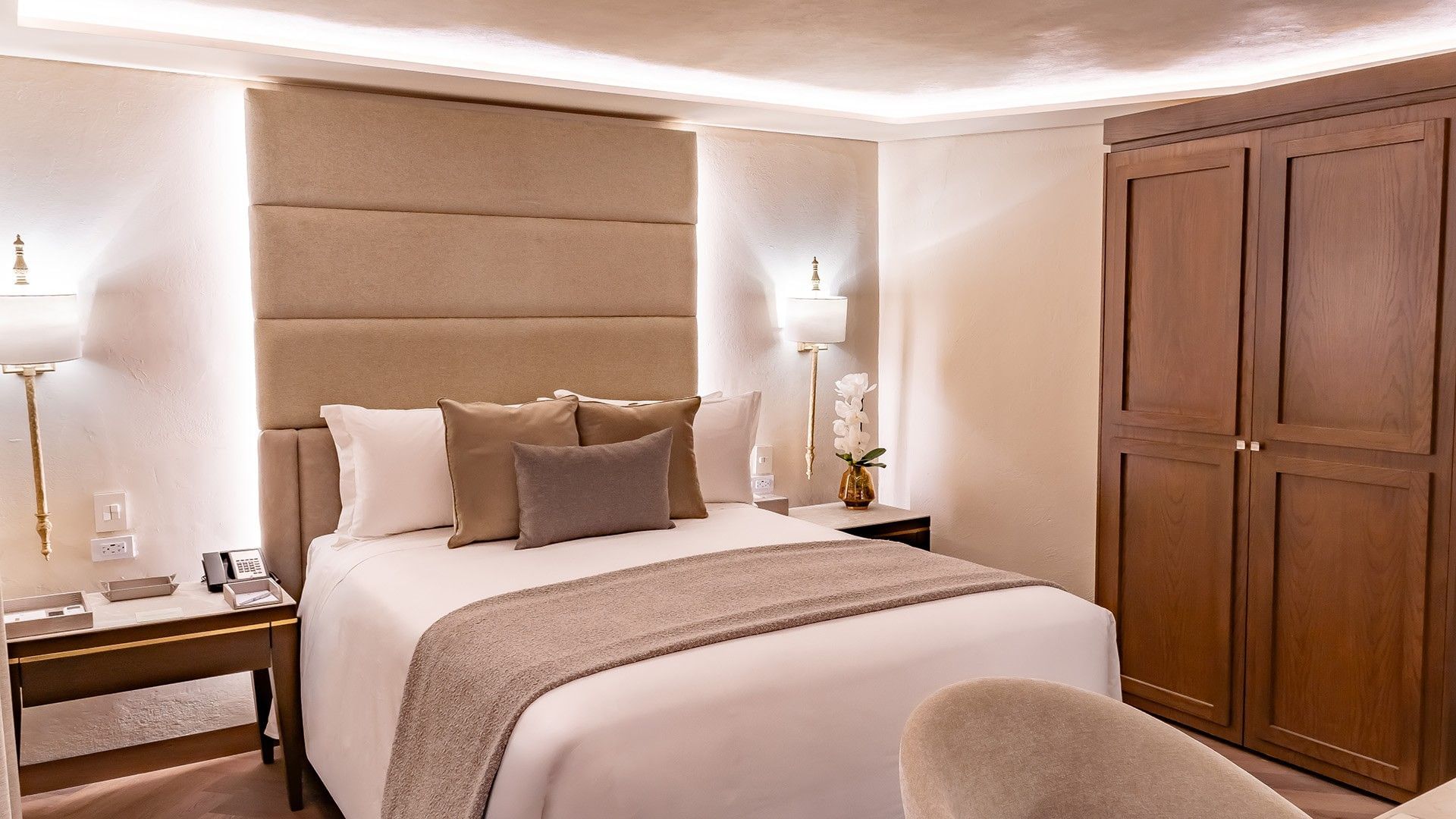 Elegant hotel bed with a tan headboard, orchid decor, and a tall wooden wardrobe in Queen Novicia at Quinta Real Puebla
