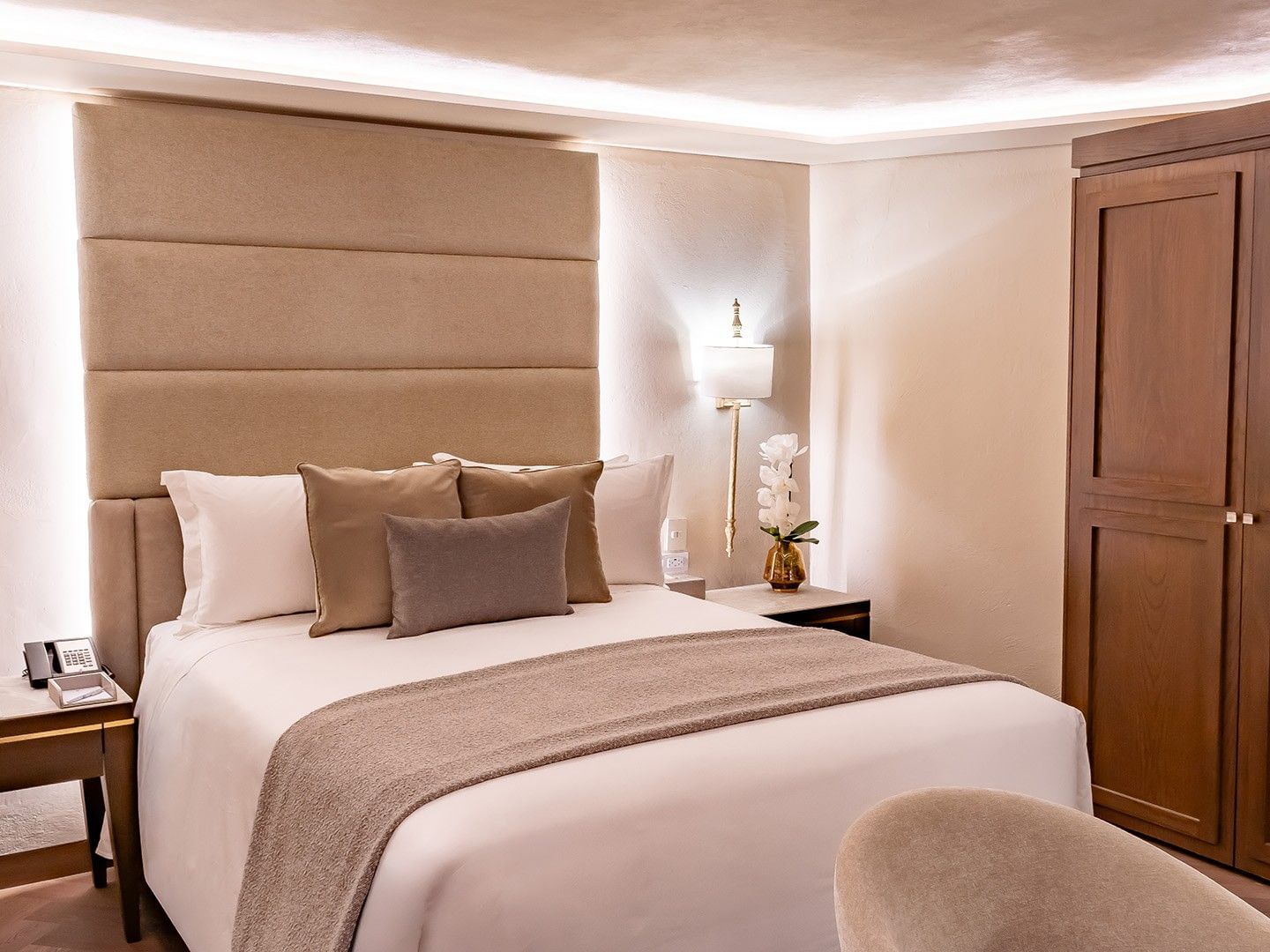 Elegant hotel bed with a tan headboard, orchid decor, and a tall wooden wardrobe in Queen Novicia at Quinta Real Puebla