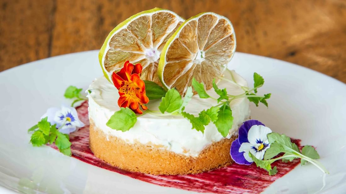 White plate with a cheesecake, topped with lime slices and flowers, sitting on a red berry sauce.