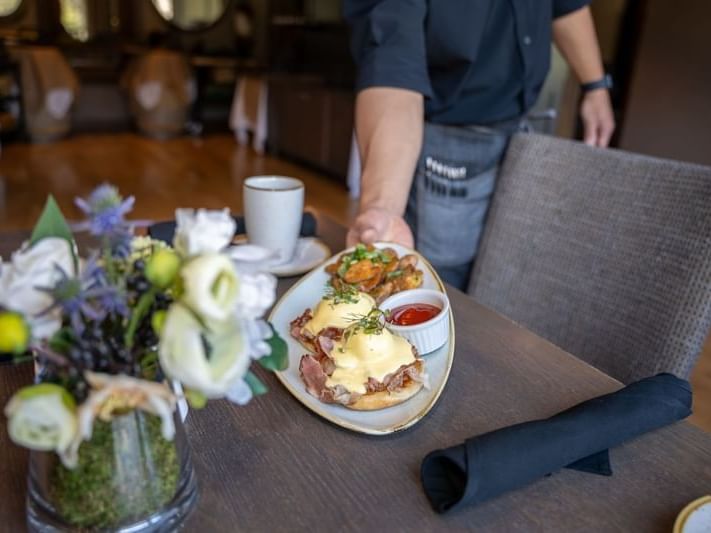 Waiter placing a plate with eggs benedict and a side dish on a table with a vase of flowers.