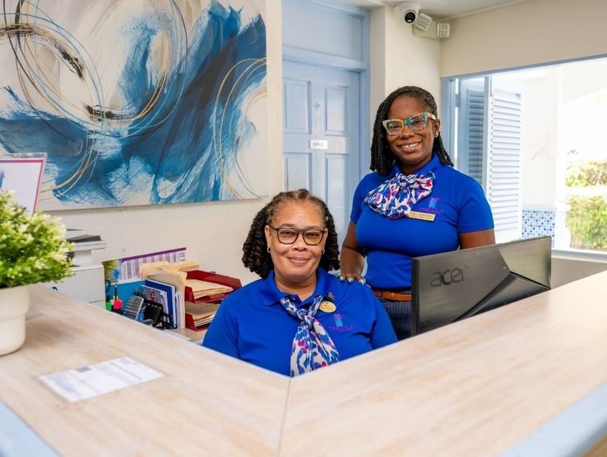 Two reception staff behind a wooden front desk with monitors and plants beneath blue abstract art at Time Out Hotel