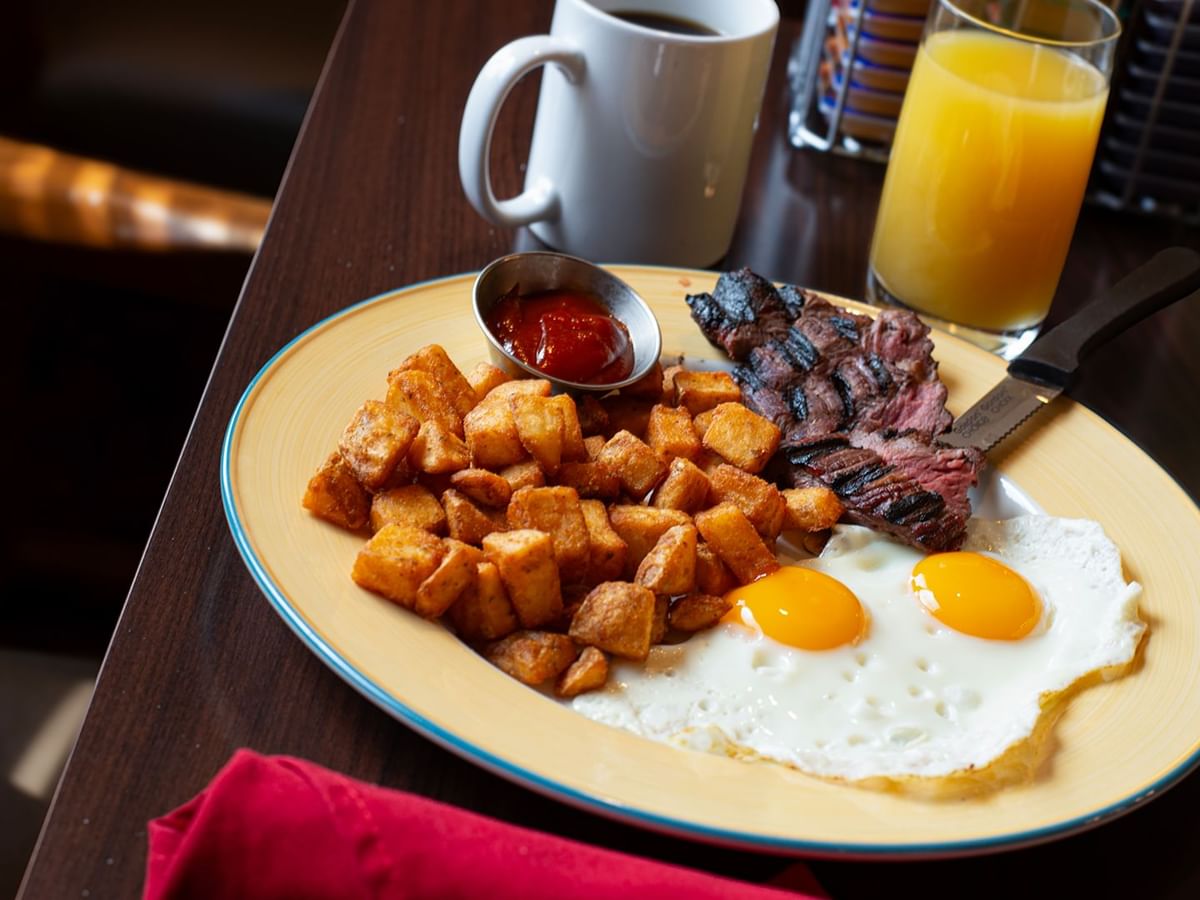 Plate of breakfast food with eggs, steak, potatoes, ketchup, coffee, and orange juice on a table.