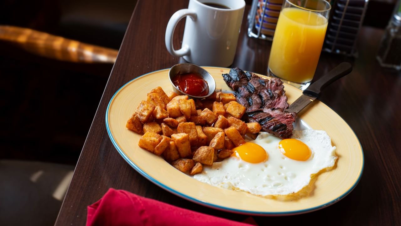 Plate of breakfast food with eggs, steak, potatoes, ketchup, coffee, and orange juice on a table.