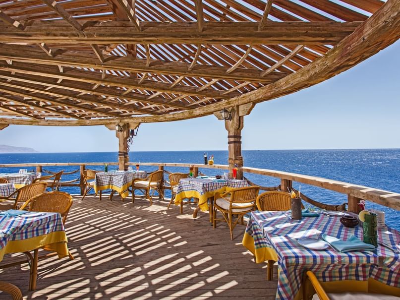 Outdoor restaurant with wooden structure, ocean view, and tables with plaid tablecloths and wicker chairs.