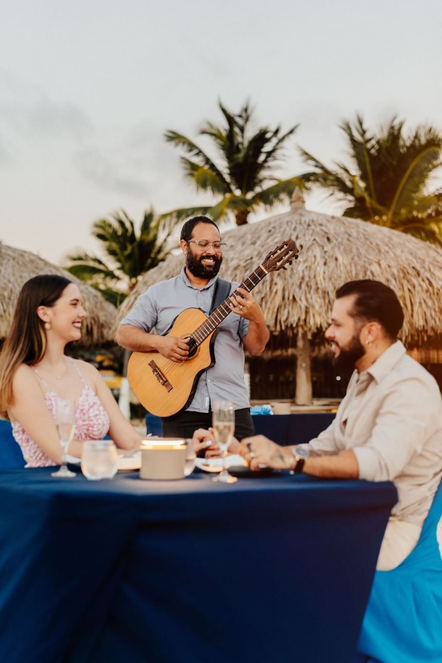 Two people seated at a table, a man playing guitar, and palm trees in the background.