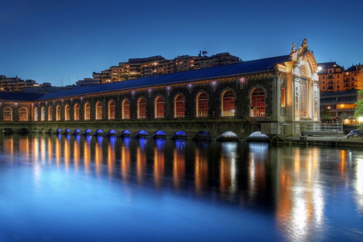 Bâtiment des Forces Motrices building with glowing arches by a blue river at night near Warwick Geneva