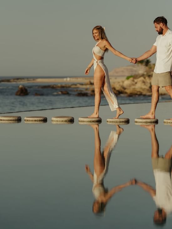 A couple holds hands walking on stepping stones across a reflective pool at Marquis Los Cabos Resort with the ocean beyond