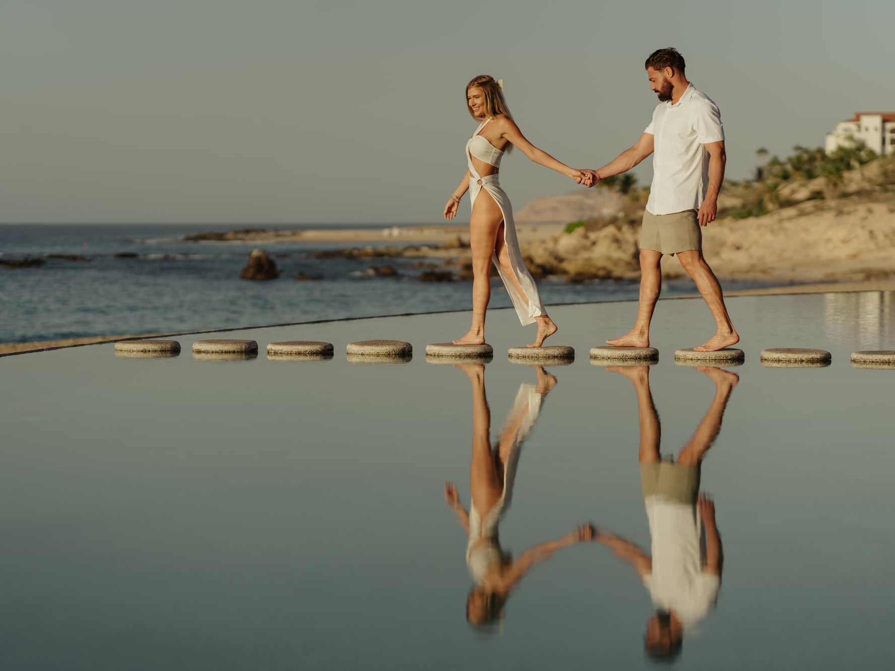 A couple holds hands walking on stepping stones across a reflective pool at Marquis Los Cabos Resort with the ocean beyond