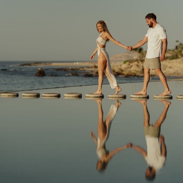 A couple holds hands walking on stepping stones across a reflective pool at Marquis Los Cabos Resort with the ocean beyond