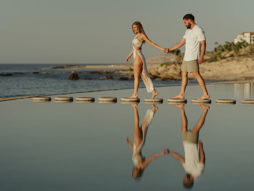 A couple holds hands walking on stepping stones across a reflective pool at Marquis Los Cabos Resort with the ocean beyond