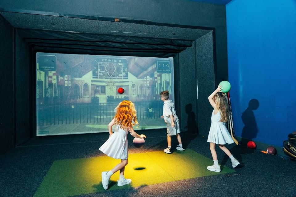 Three kids playing a sports simulator game facing a large projection screen on green turf at Hotel X Toronto