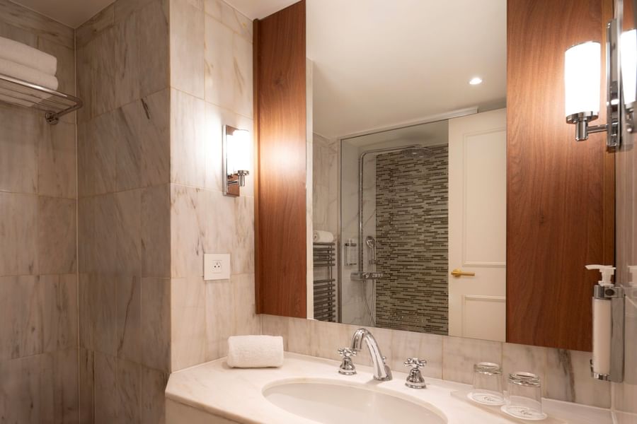 Modern bathroom with marble vanity, white sink, wall-mounted mirror, and glass-enclosed shower at Warwick Brussels Grand-Place.