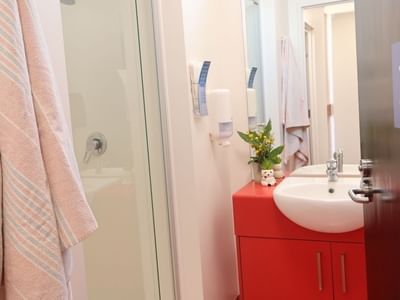 Pink towel hanging on glass shower door with sink and red cabinet below.