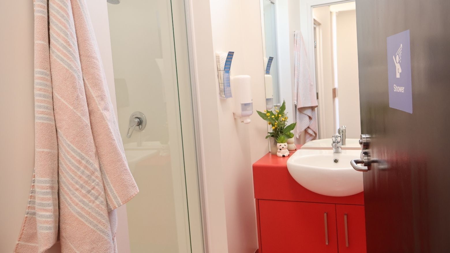 Pink towel hanging on glass shower door with sink and red cabinet below.