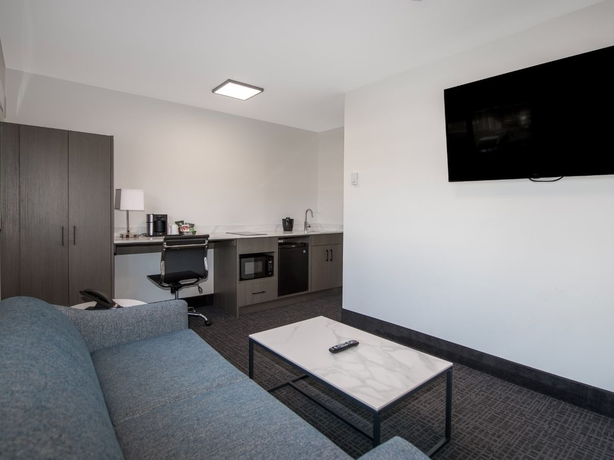 A modern hotel suite with a couch, coffee table, flat-screen TV, and a small kitchenette.