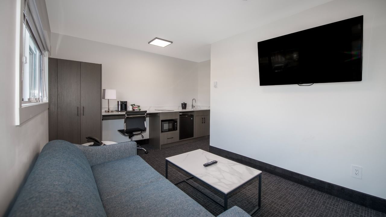 A modern hotel suite with a couch, coffee table, flat-screen TV, and a small kitchenette.