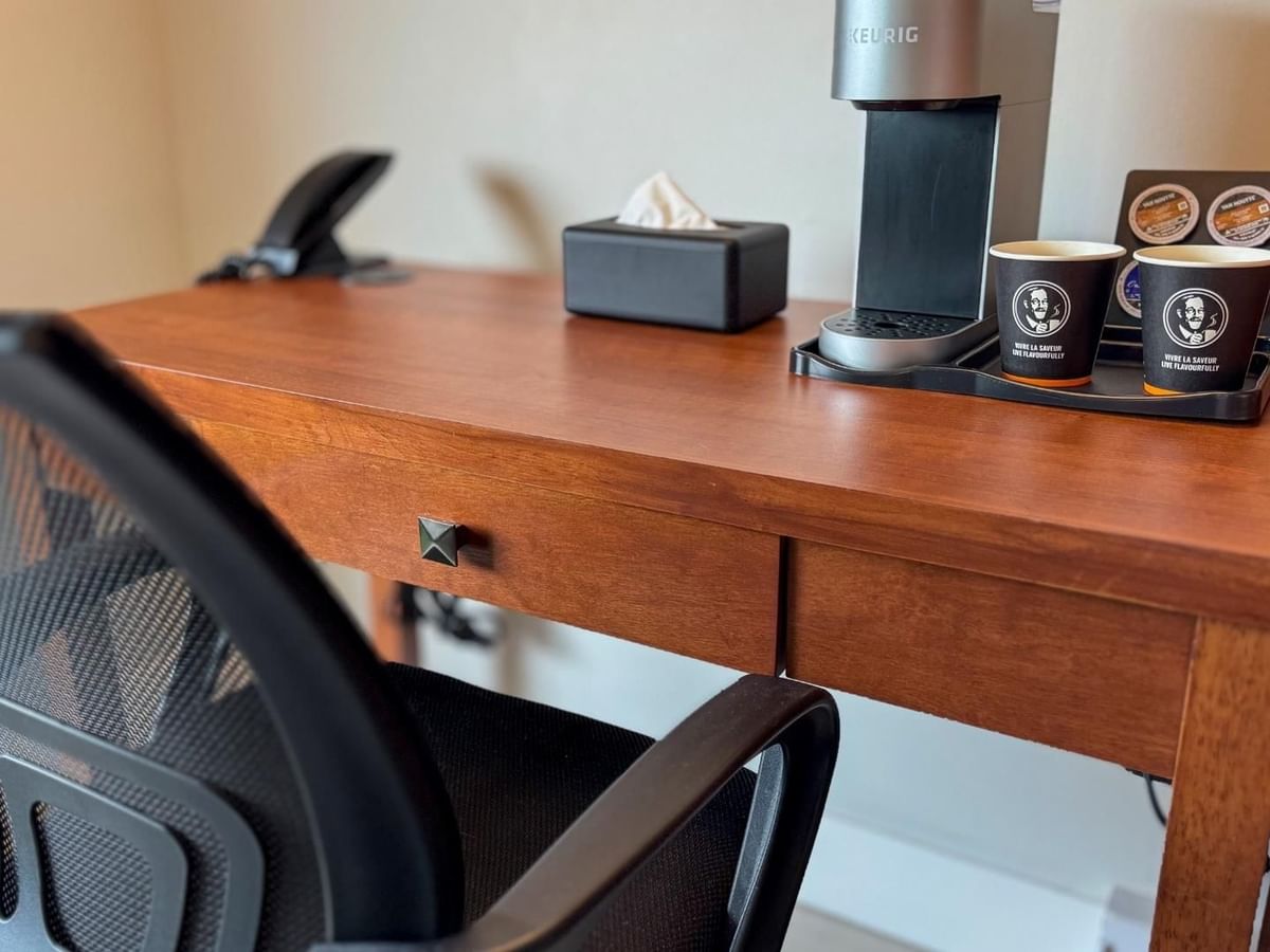 Wooden desk with chair, coffee maker, two cups, tissue box, and phone on top.