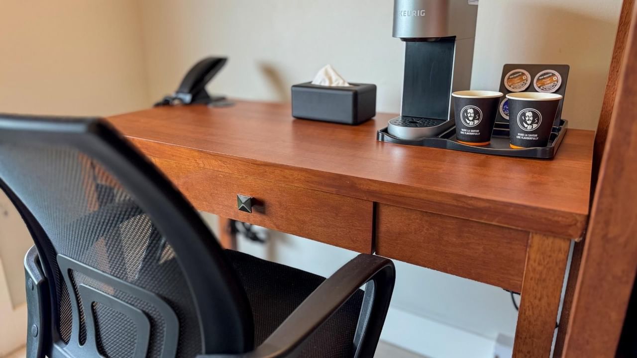 Wooden desk with coffee maker, two cups, tissue box, phone and a black mesh chair.