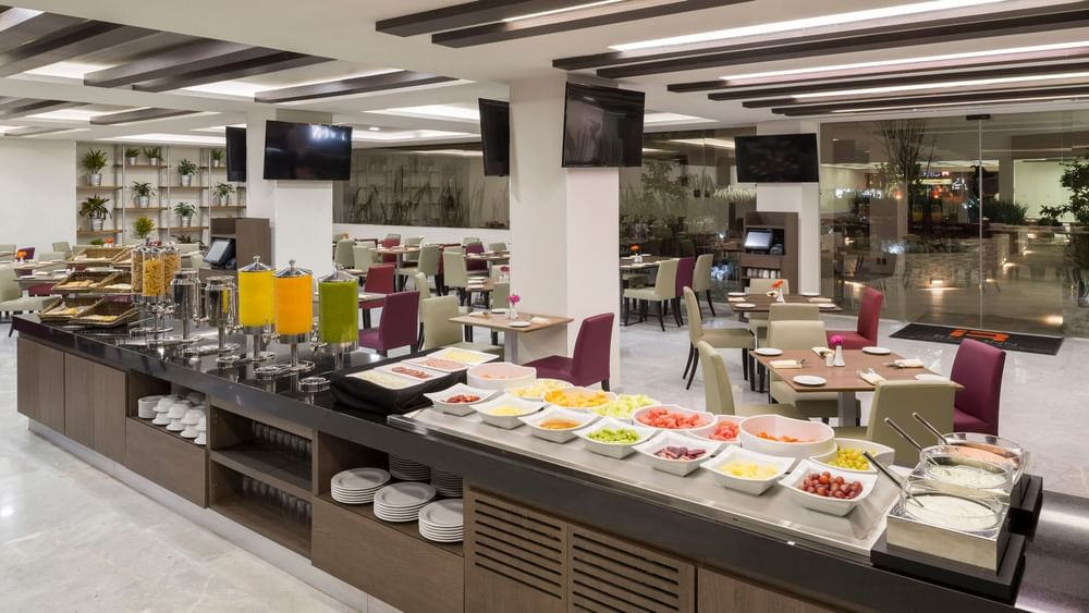 Bright dining area in Stock Cafe with a breakfast bar, fruit selection, and purple upholstered chairs at Real Inn Celaya