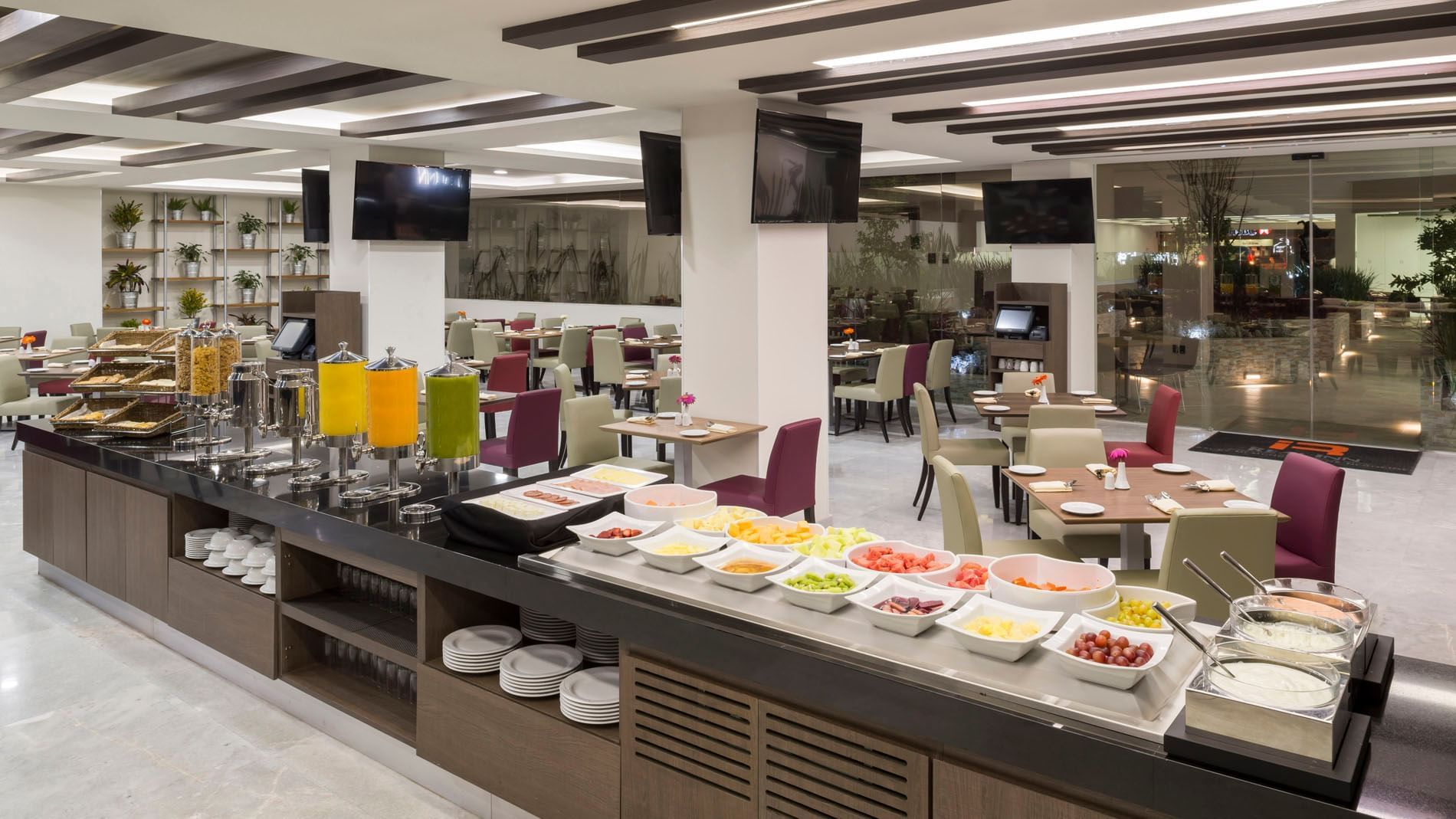 Bright dining area in Stock Cafe with a breakfast bar, fruit selection, and purple upholstered chairs at Real Inn Celaya